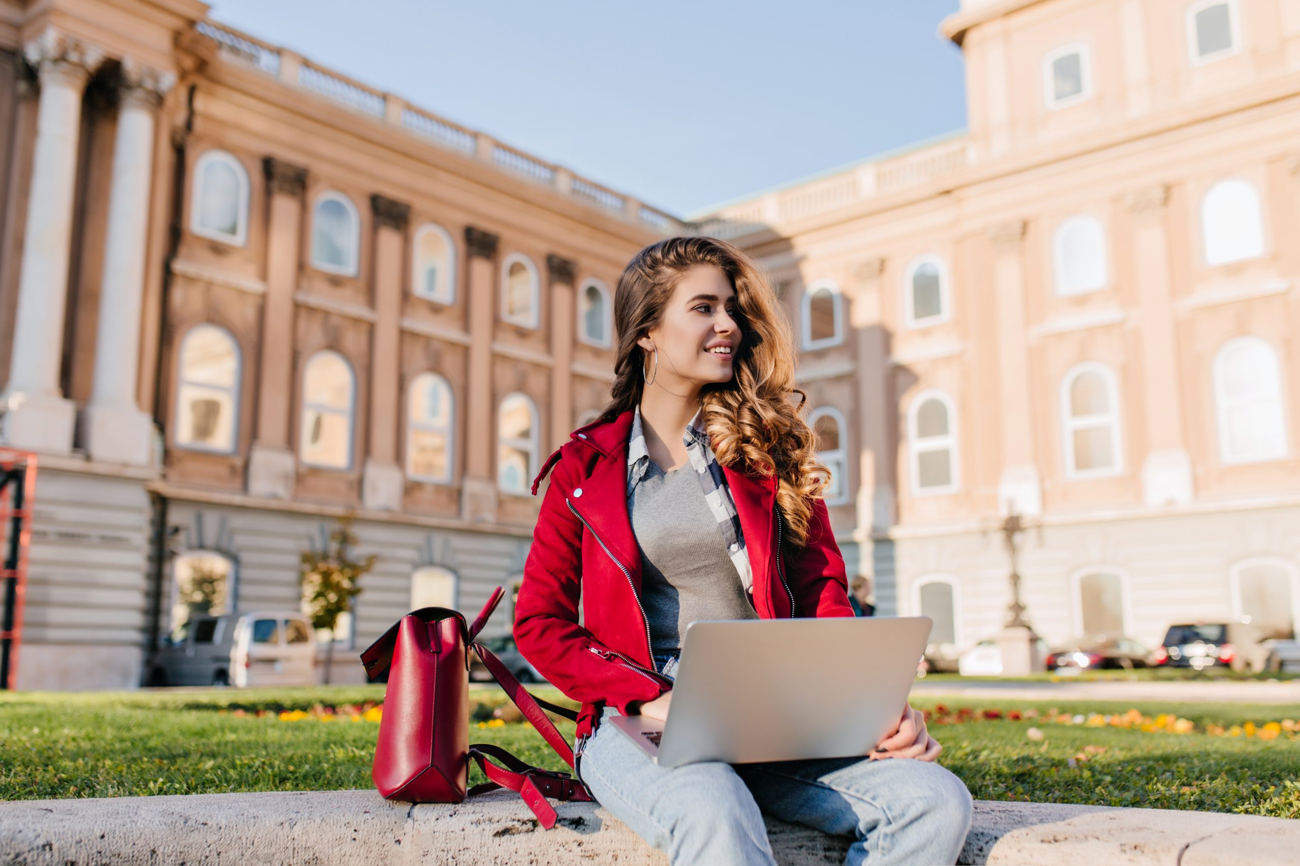 Interested dark-haired girl wears casual attire chilling in park near university and using laptop. Outdoor photo of fashionable female freelancer working with computer on the grass..