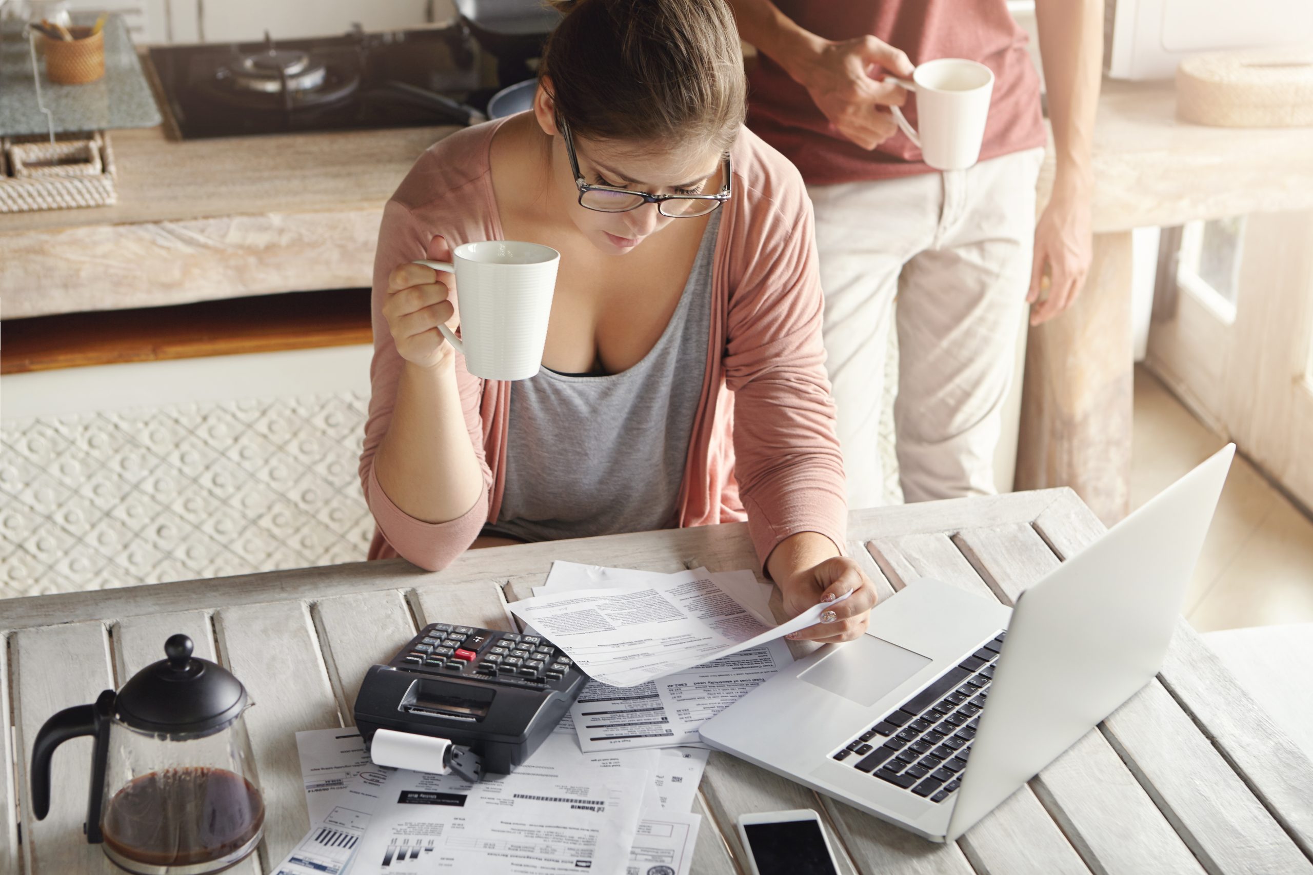 Top view of serious young woman wearing glasses managing family budget, calculating domestic expenses, paying utility bills online, using laptop pc and calculator while drinking coffee in kitchen